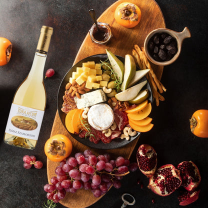 Holzbrett mit Käsewürfeln, verschiedenen Käsesorten, Nüssen, Birnenspalten, Trauben, Persimonen, Granatapfelhälften und einem Glas Marmelade daneben eine Flasche Tokajicum Száraz Szamorodni.