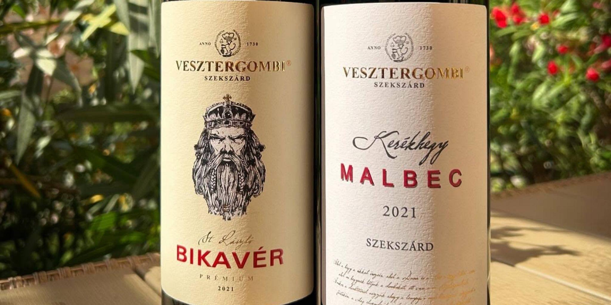 Zwei Weinflaschen mit Etiketten: links 'VESZTERGOMBI BIKAVÉR PREMIUM', rechts 'VESZTERGOMBI Kékfrankos MALBEC 2021 SZEKSZÁRD' vor unscharfem Hintergrund mit grünen Blättern und roten Beeren.