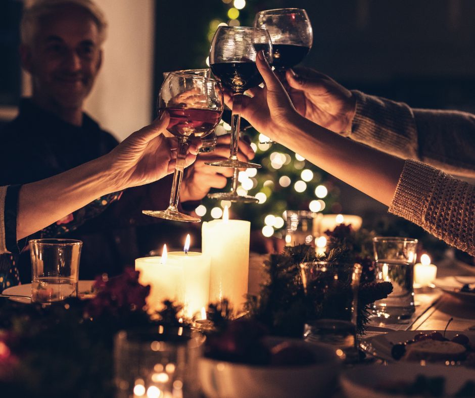 Fünf Gläser mit Wein werden angestoßen, umgeben von Kerzen und festlicher Dekoration auf einem Tisch. Im Hintergrund sind Lichter eines Weihnachtsbaums sichtbar.
